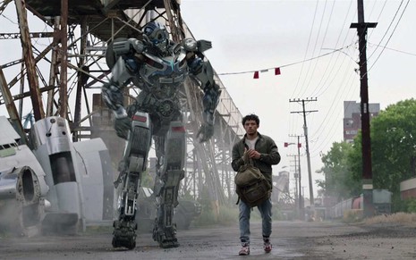 Mirage e Noah Diaz caminham lado a lado com uma ponte velha ao fundo em cena de Transformers