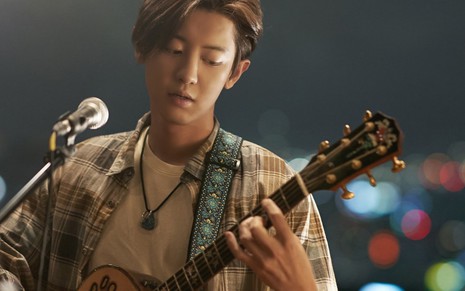 Park Chanyeol veste uma camiza xadrez, um colar, enquanto toca violão e canta
