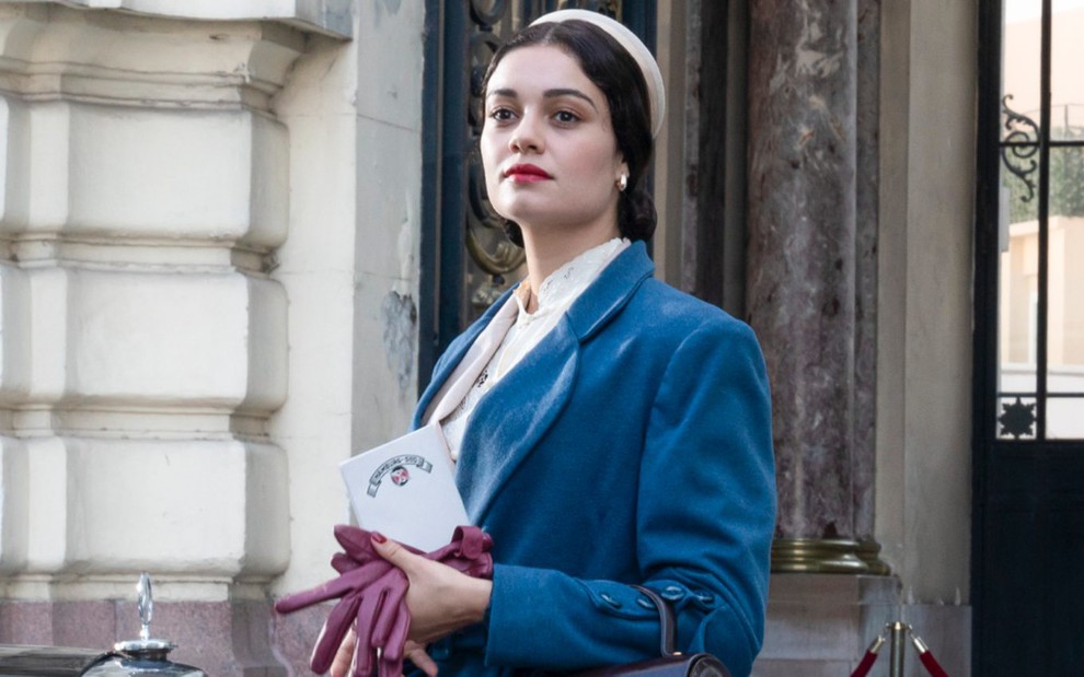 Sophie Charlotte de casaco azul e luvas roxas nas mãos, olha para o horizonte caracterizada como sua personagem em Passaporte para Liberdade
