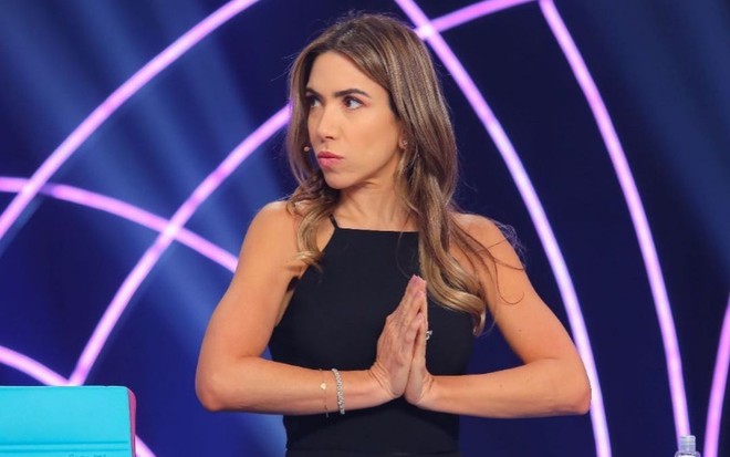 Patricia Abravanel no estúdio do Roda a Roda Jequiti com as duas mãos unidas em sinal de oração. Seu rosto está de perfil com uma expressão de preocupação.