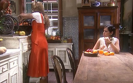 As atrizes Patricia Pillar e Gloria Pires como Luana e Marieta/Rafaela em O Rei do Gado; a primeira está em pé, de costas para a segunda enquanto mexe com comida e a segunda está sentada, comendo, e olhando para o lado enquanto conversa