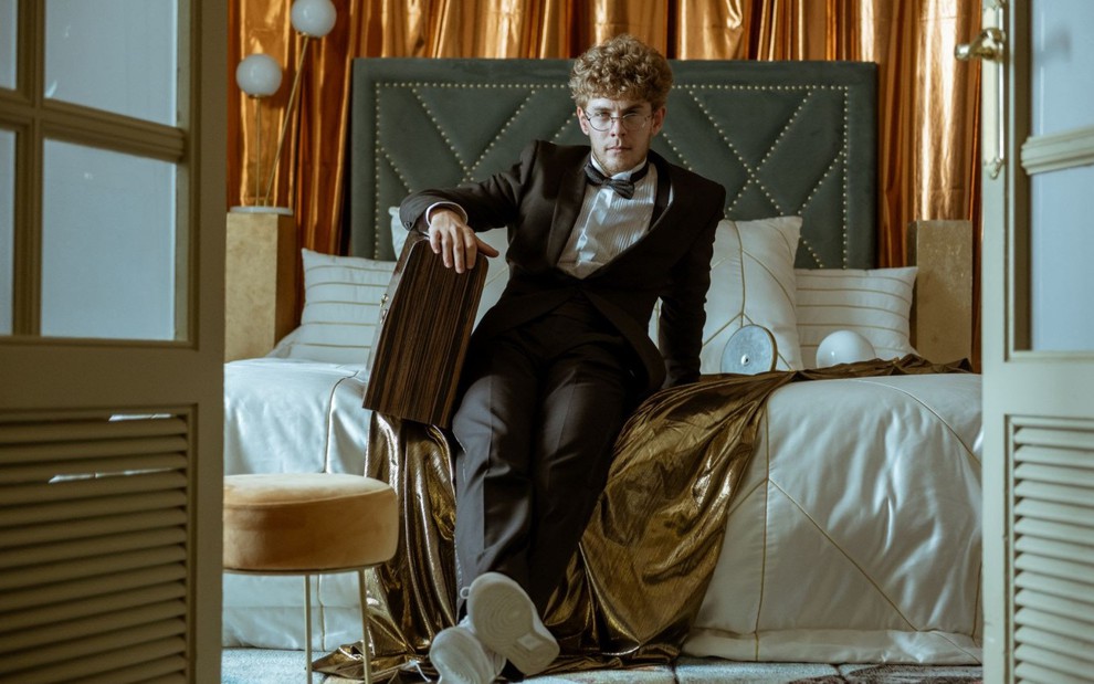 Patrick Criado em cena de La Casa de Papel; ele posa em um quarto e está sentado na cama