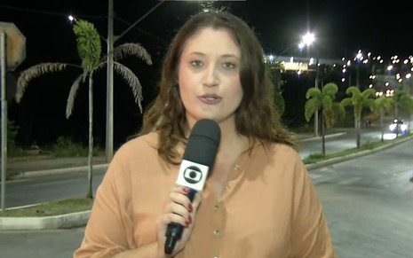 Paula Alves em frente a uma rodovia, segurando o microfone da Globo
