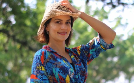 Paula Barbosa coloca a mão no chapéu de palha que usa em um cenário cheio de árvores