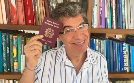 O ator Pualo Betti de camisa listrada posando em seu seu Instagram com um passaporte europeu levatado pela mão direita e próximo ao rosto. O ator está sorrindo e no findo é possível observar uma estante repleta de livros.