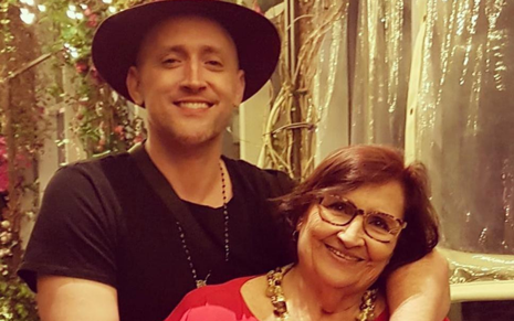 Paulo Gustavo usa camiseta preta e chapéu e abraça sua mãe, Déia Lúcia, que usa vestido vermelho