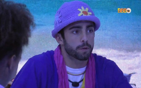 Pedro Scooby no Almoço do Líder dentro da casa do BBB 22, ele veste um chapéu de crochê florido e uma camisa azul com echarpe rosa no pescoço