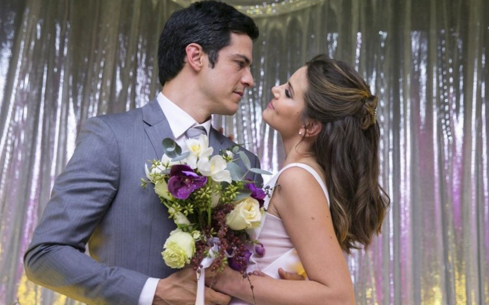 Eric (Mateus Solano) e Luiza (Camila Queiroz) estão vestidos de noivos e abraçados, ela segura um buquê de rosas brancas na mão em cena de casamento em Pega Pega
