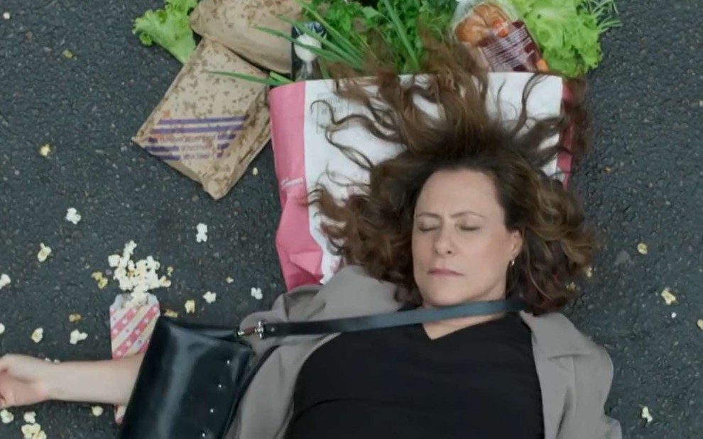 Atriz Elizabeth Savala surge deitada no meio da rua com compras ao fundo em cena de Pega Pega