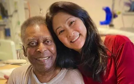 Pelé está sentado na cama do hospital, sorrindo; Márcia Aoki está apoiada nele, também sorrindo