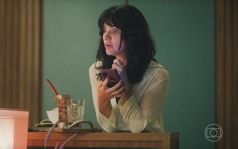 Em cena de Terra e Paixão, Debora Ozorio está com a expressão de espanto, segurando um aparelho de celular
