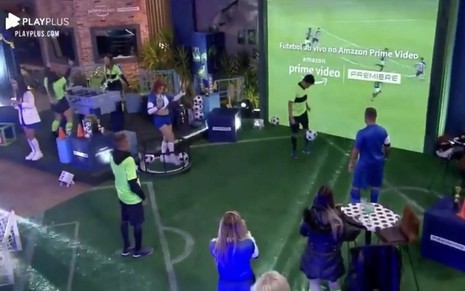 Participantes do Power Couple Brasil 5 vestidos com roupas azuis de jogadores de futebol e jogando com uma bola. O chão está com um tapete que parece um gramado e gols foram colocados na casa do programa. Um telão grande mostra o logo do Premiere.