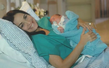 Regina Duarte está deitada numa cama de hospital e segura um bebê em cena como Helena na novela Por Amor (1997)