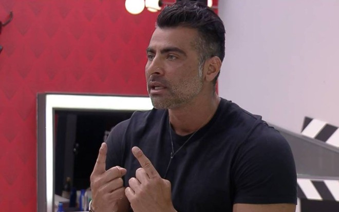 JP olha para o lado, veste camiseta preta e faz sinal com os dois dedos indicadores das mãos; o modelo está na frente de uma parede branca e outra vermelha que tem um espelho