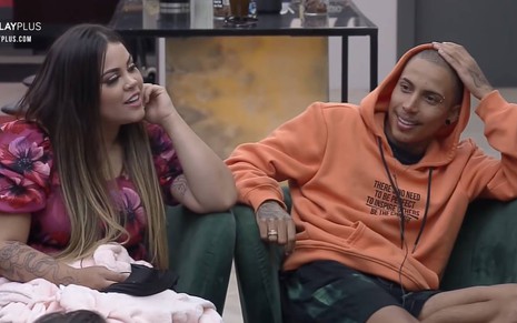 Mari está sentada ao lado de Matheus, ela usa vestido rosa e está com uma mão na boca; Matheus está sentado ao lado de Mari, está com uma mão na cabeça e usa blusa laranja