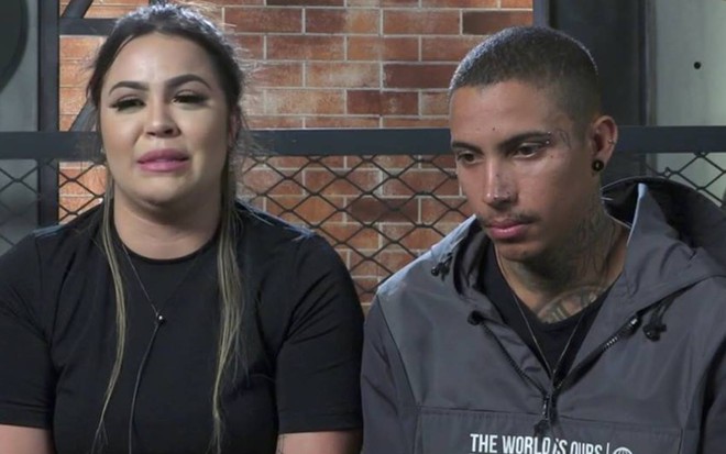 Mari está sentada ao lado de Matheus, ela usa camiseta preta e está chorando; Matheus olha para baixo e usa blusa cinza