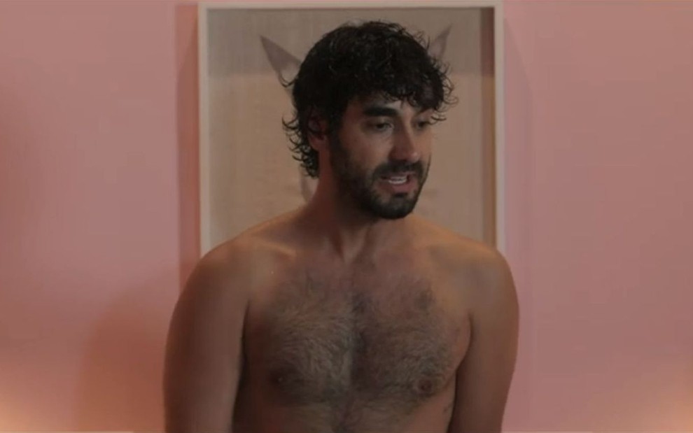 Gabriel Godoy está sem camisa em frente a um quadro e a uma parede cor de rosa em cena de Desjuntados