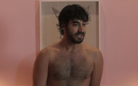 Gabriel Godoy está sem camisa em frente a um quadro e a uma parede cor de rosa em cena de Desjuntados