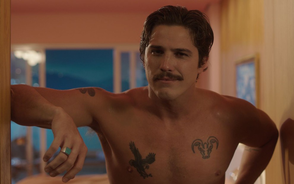Romulo Arantes Neto está sem camisa, com o braço apoiado no vão da porta, com um bigode de safadinho e tatuagens no peitoral