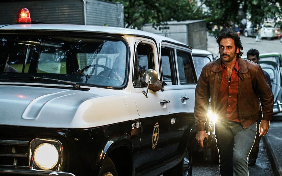 Com jaqueta de couro, Flavio Tolezani segura uma arma e caminha ao lado de uma viatura da Polícia Civil em cena da série Dom