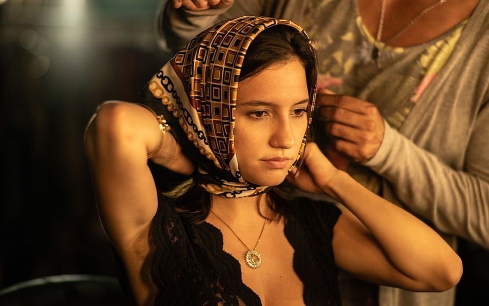 Lorena Comparato olha séria para frente, está com um lenço na cabeça sendo ajeitado por outra mulher