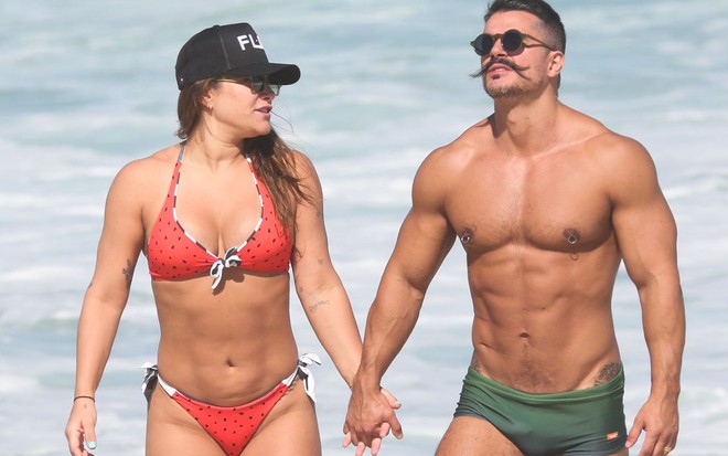 Priscila Fantin, de biquíni vermelho, caminha de mãos dadas com o marido, Bruno Lopes, que usa sunga