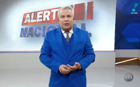 Sikêra Jr. de terno azul com as mãos juntas na altura da cintura com telão ao fundo no programa Alerta Nacional, da RedeTV!