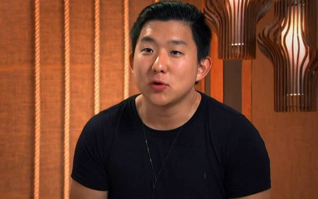 Pyong está sentado, veste camiseta preta e está na frente de uma parede de madeira