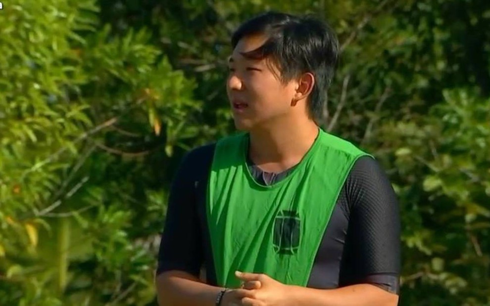 Pyong olha para o lado, ele veste camiseta preta e colete verde