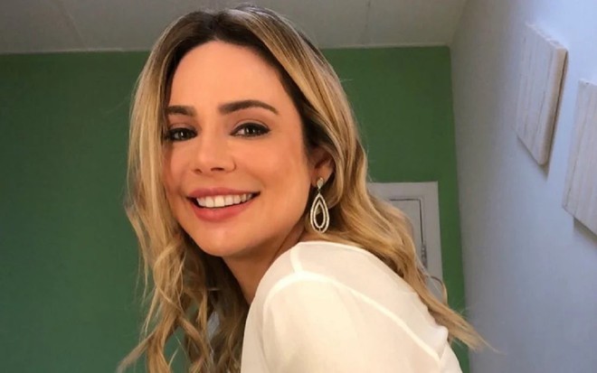 A jornalista Rachel Sheherazade sorrindo, com parede verde no plano de fundo, de camisa branca