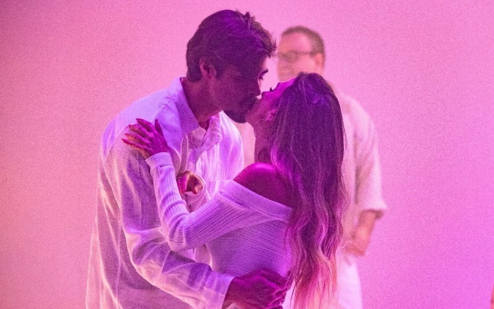 Em fundo rosa, Rafa Vitti beija Tatá Werneck; ambos vestem roupa branca. Ao fundo aparece o comentarista Everaldo Marques
