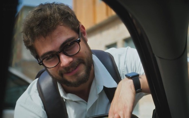 Em cena de Terra e Paixão, Rafael Vitti usa mochila e óculos e está sorrindo para alguém dentro de um carro