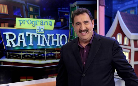 Ratinho usa um terno preto com uma camisa preta, e olha para a câmera sorridente no cenário de seu programa na emissora de Silvio Santos