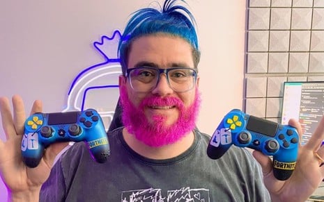 Raulino de Oliveira Maciel, o RaulZito, com o cabelo azul e a barba rosa, segurando dois controles de videogames