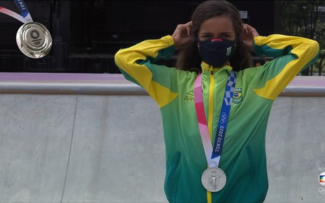 Rayssa Leal, a Fadinha, usa um casaco verde e amarelo, e uma máscara preta. Ela está com o cabeço amarrado e a medalha de prata no peito.