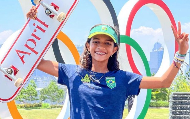 Rayssa Leal posando em frente aos arcos olímpicos para o Instagram durante sua passagem pelos Jogos Olímpicos de Tóquio. Ela veste uma camisa azul, uniforme oficial do Brasil, e uma viseira verde. A skatista está soorrindo segurando o skate pro alto.