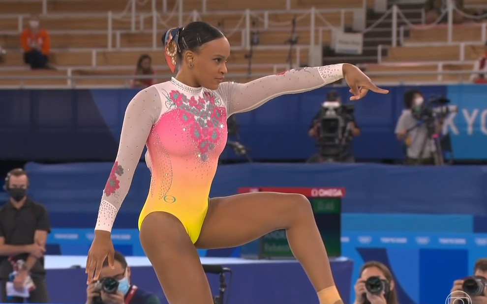 Rebeca Andrade com um collant rosa, amarelo e branco, apontando para frente e ajoelhada durante apresentação nos Jogos Olímpicos de Tóquio