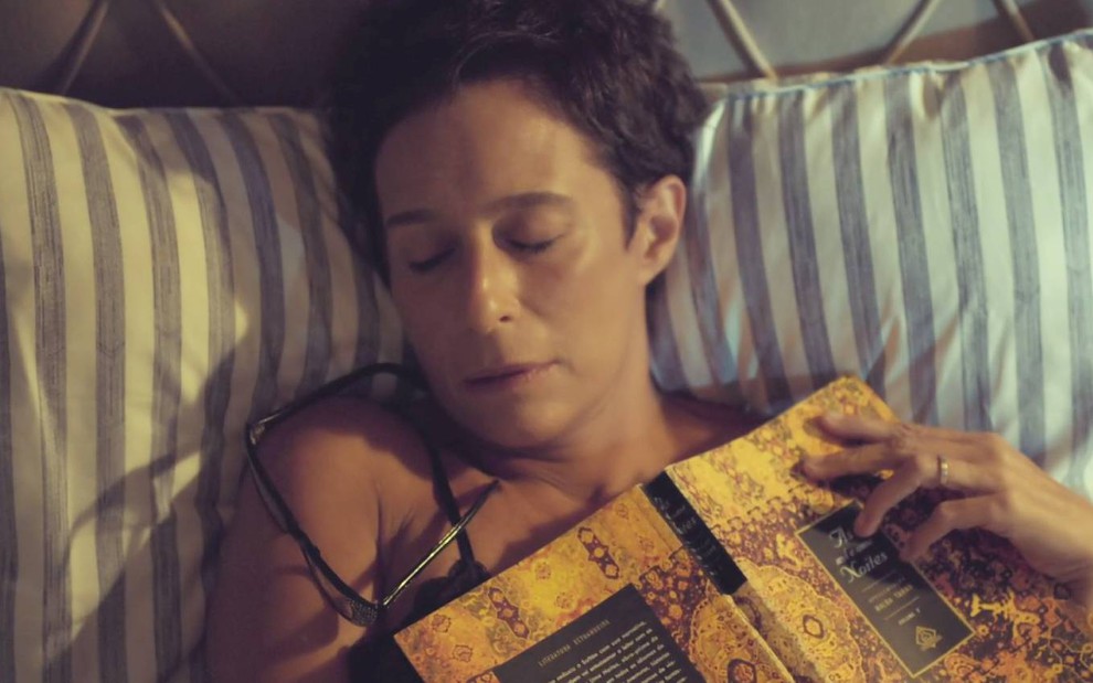 Atriz Andrea Beltrão surge de olhos fechado e deitada em cama, enquanto segura livro, em cena de Um Lugar ao Sol