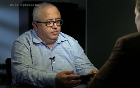 Ricardo Rocha está sentado, usa camisa azul clara e óculos, e entrevista exibida pelo Domingo Espetacular, da Record