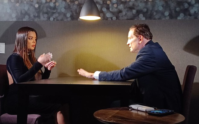 Ingrid Lana conversa com Roberto Cabrini em uma mesa mal iluminada; os dois têm expressões sérias