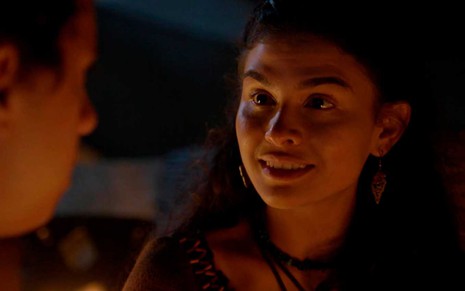 A atriz Allana Lopes está caracterizada como Bila com o rosto iluminado pela luz emanada por uma fogueira em cena noturna de gÊNEEIS