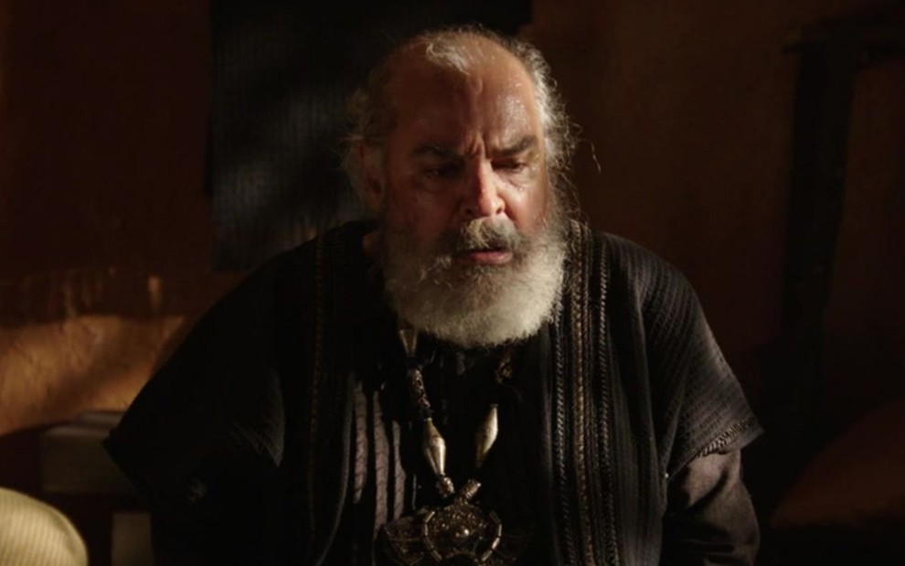 Roberto Bomfim caracterizado como Betuel em cena de Gênesis: ator está sentado e olha com desespero para baixo