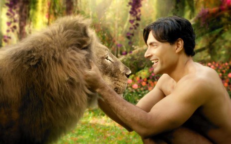O ator Carlo Porto como Adão interage com um leão, criado por efeitos especiais, em um cenário paradisíaco como um folheto de Testemunha de Jeová em cena de Gênesis