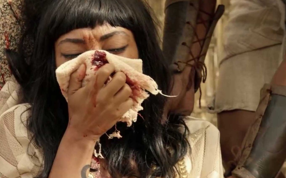 Com expressão de dor, a atriz Dandara Albuquerque como Neferíades leva um lenço à frente do rosto, e o tecido está manchado de sangue, em cena de Gênesis