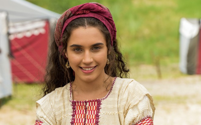 Giovanna Coimbra posa sorridente, com lenço rosa no cabelo e vestido bege e vermelho como Diná de Gênesis