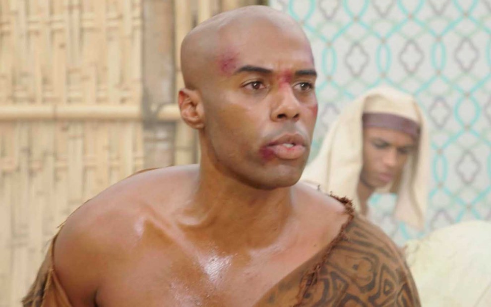 O ator Dudu de Oliveira como o Abumani com a roupa rasgada, sangue no canto da boca e escoriações em cena de Gênesis