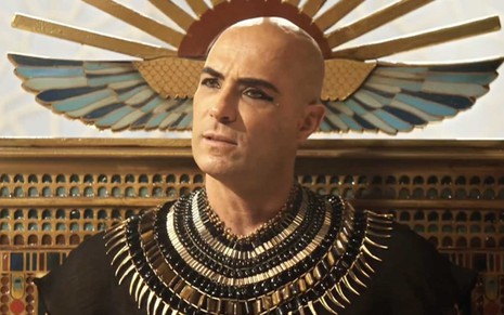 Sentado em um trono, o ator Fernando Pavão está careca e com os olhos marcados de kajal como o faraó Sheshi em cena de Gênesis