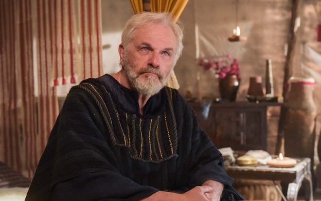 O ator Henrique Pagnoncelli caracterizado como Isaque, com barba e cabelos brancos, e olhos esbranquiçados como se fosse cego, em cena de Gênesis