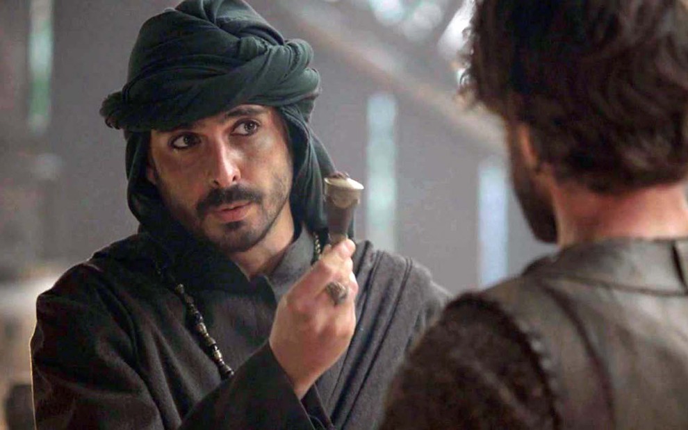 O ator Iano Salomão segura um vidro de veneno com a mão direita e oferece a um homem, que está de costas, como o Ismael em cena de Gênesis