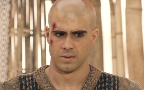 Juliano Laham em cena de Gênesis: ator está com o rosto machucado, cabelo raspado e túnica marrom